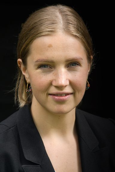 Portret van een jonge vrouw met een lichte huid en blond haar, in een zwarte blazer.